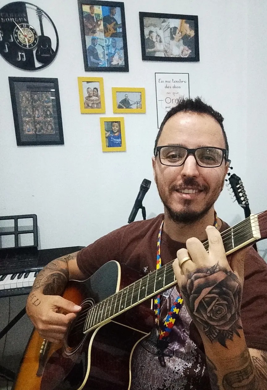 Carlos Borges sorrindo com o violão na sala de aula; atrás dele, a parede branca com o relógio de vinil e as molduras amarelas